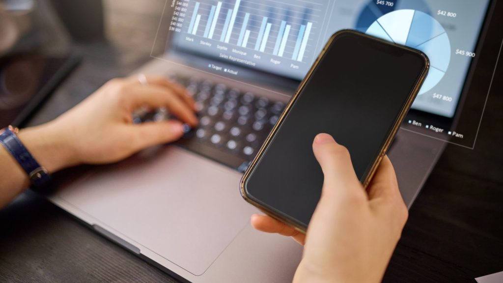 Hand holding a smartphone over a laptop showing analytics charts