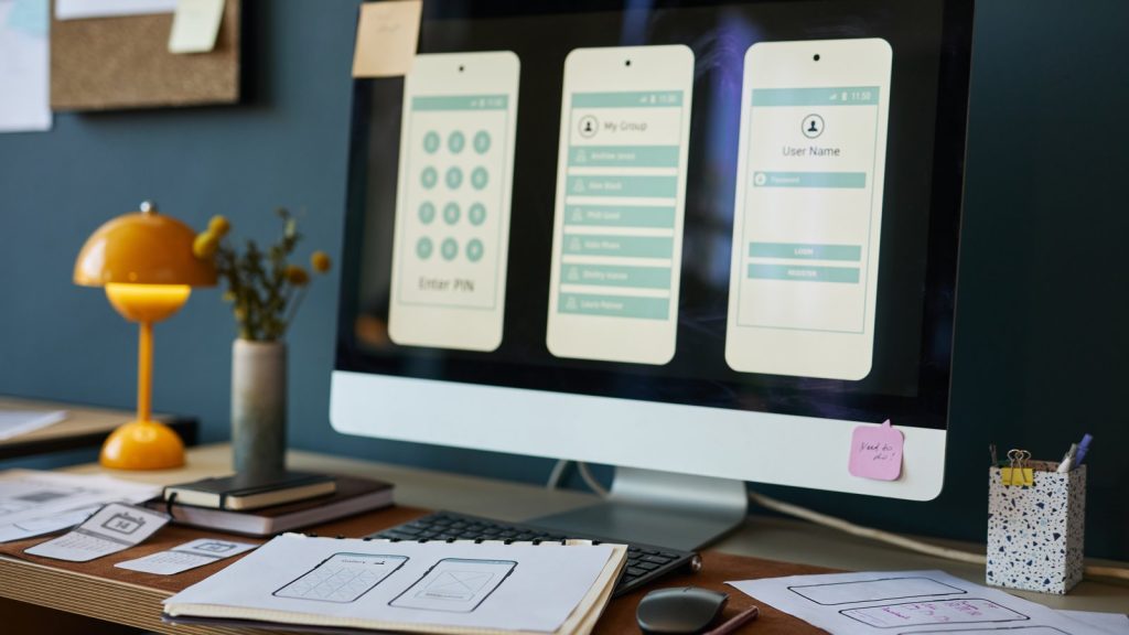 Mobile app screen designs on a computer monitor at a desk with a lamp