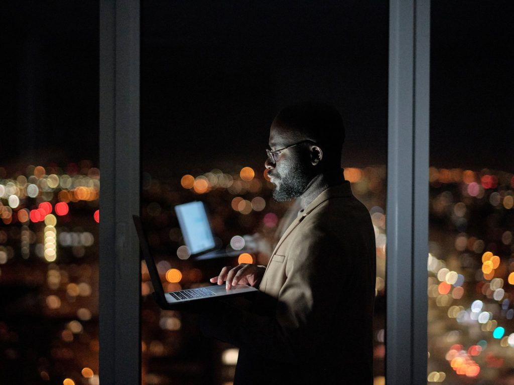 Man working on a laptop late at night by a window with city lights